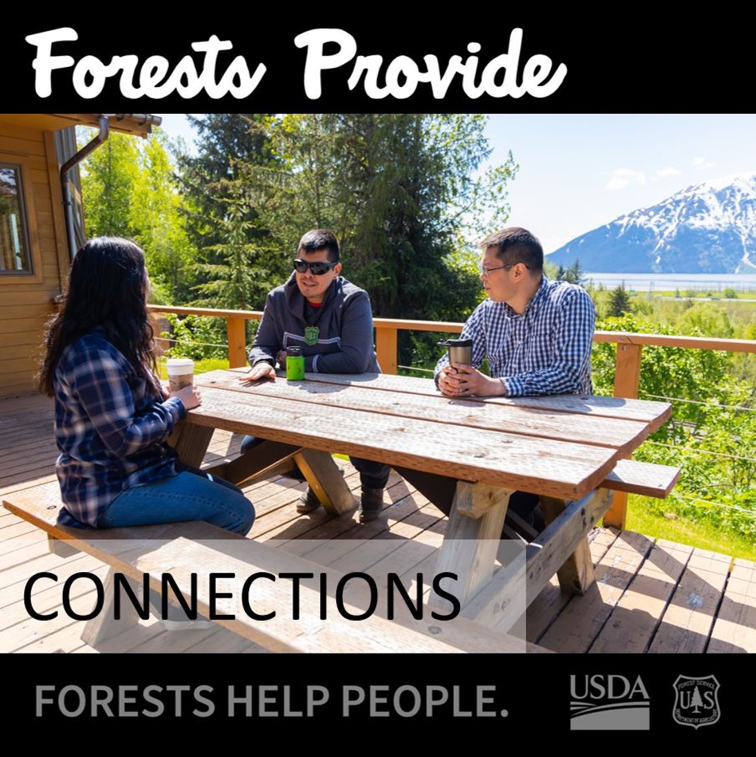 Three people sitting at a picnic table on the wood deck of a remote cabin. A body of water and snow capped mountain loom in the far background.