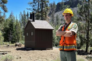 A man wearing a USDA Forest Service uniform, yellow hard hat, and bright orange safety vest, arms folded, standing near a forest restroom.