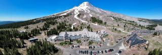 An aerial view of Timberline Lodge with a partially snow covered mountain peak in the direct background and a clear blue sky.